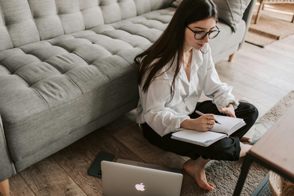 Woman journaling on the floor with a laptop nearby during a chronic pain therapy session, representing chronic pain obsessions and emotional processing