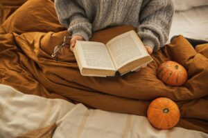 Person reading in bed with cozy fall blankets and pumpkins, symbolizing seasonal changes that affect sleep