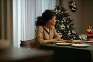 A woman sitting at a holiday dinner table looking away thoughtfully, with a Christmas tree in the background — representing the emotional strain and pain flares often experienced when living with chronic pain during the holidays.