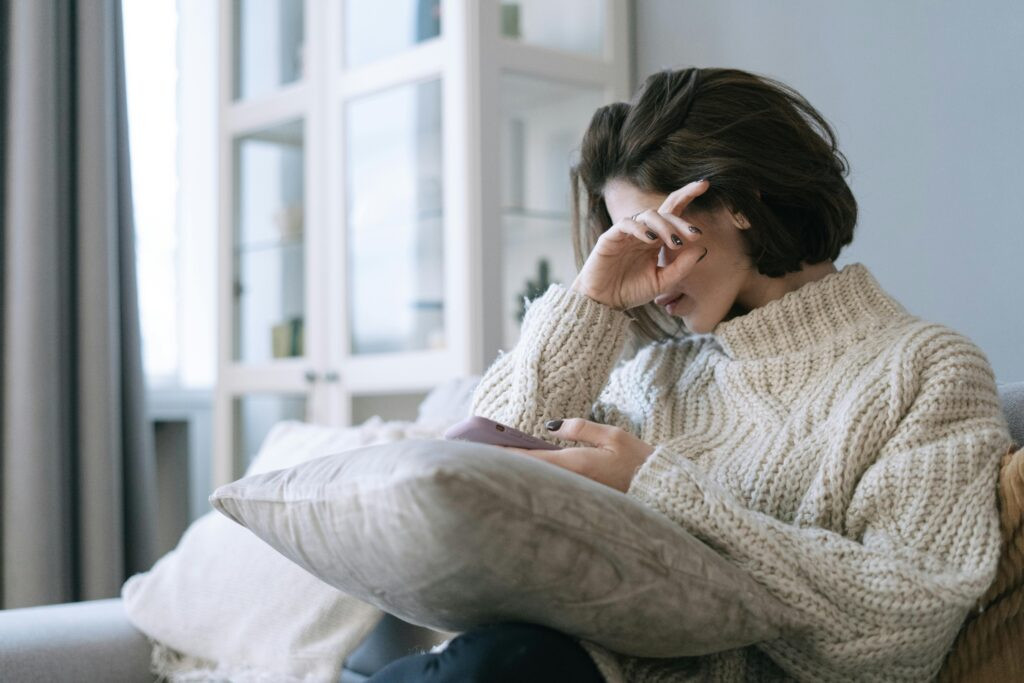 Woman sitting on a couch holding her phone while experiencing aching joints from cold weather, representing how cold weather can make chronic pain feel worse in winter.